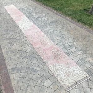 Puede incluir: Un largo corredor de alfombra rectangular de color rosa y crema con un patrón floral. La alfombra está colocada sobre un camino de concreto gris.