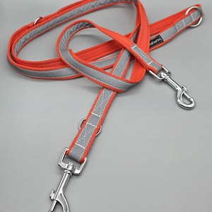 May include: An orange dog lead with reflective grey trim and silver metal clasps and rings. The lead is made of a durable material and is designed for pet safety during walks. The lead is shown against a grey background.