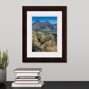 May include: A framed photograph of a mountain range with a blue sky and white clouds. The mountain range is in the distance and there are bushes in the foreground.