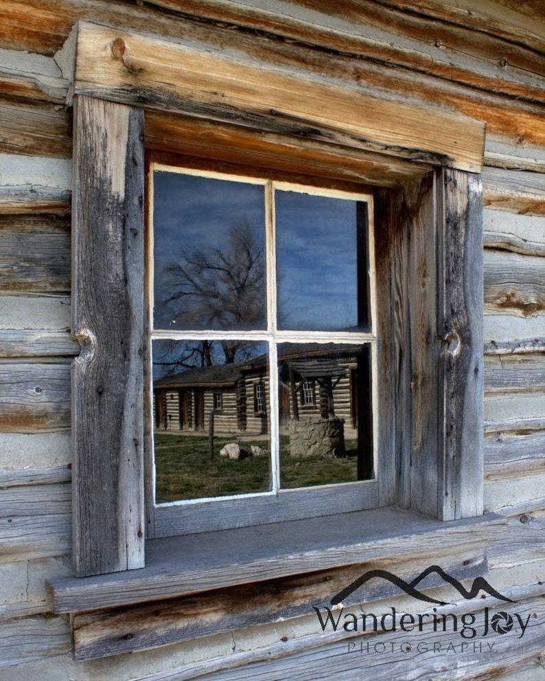 K&ouml;nnte beinhalten: Ein verwittertes Holzfenster mit Blick auf ein Geb&auml;ude, das sich in den Glasscheiben spiegelt. Das Fenster ist von einem dicken Holzrahmen umgeben und hat eine einfache wei&szlig;e Verkleidung.