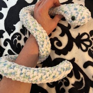 May include: A soft, crocheted snake toy in white with blue and green flecks. The snake has a rounded head with black button eyes. The cuddly toy is coiled and appears to be handmade.