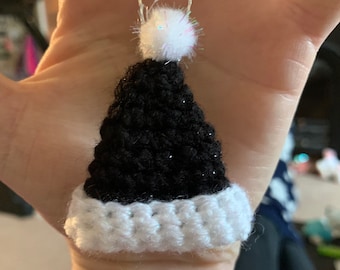 May include: A black and white crocheted Santa hat pendant with a silver chain. The hat has a white brim and a fluffy white pom-pom at the top. The pendant is a festive holiday accessory.