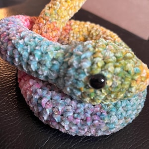 Op de afbeelding: Een gehaakt regenboog slang speelgoed met een zwart oog. De slang is opgerold in een cirkel.