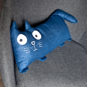 Puede incluir: Una almohada azul con forma de gato con ojos, nariz y bigotes bordados en blanco. La almohada está sobre un sillón gris.
