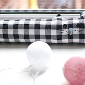 Può includere: Un fermaporta a forma di gatto a scacchi neri e bianchi con il naso blu e i baffi. Il fermaporta è su una superficie bianca con luci a sfera di cotone bianche e rosa.