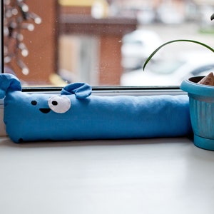 Può includere: Un fermaporta in tessuto blu a forma di cane dei cartoni animati con un occhio bianco e una bocca nera. Il fermaporta è seduto su un davanzale bianco accanto a un vaso blu con una pianta verde.