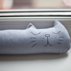 Op de afbeelding: Een lichtblauwe stoffen deurstop in de vorm van een kat met een zwart geborduurd gezicht. De kat heeft gesloten ogen en een kleine neus.