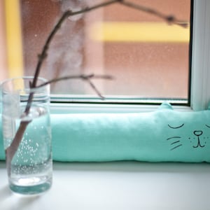 以下が含まれることがあります： 黒い目とひげのある猫の形をしたターコイズブルーのすきま風ストッパー。すきま風ストッパーは白い窓枠の上に置かれています