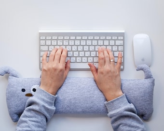 May include: A person's hands are typing on a silver and white wireless keyboard. A gray wrist rest shaped like a dog is positioned in front of the keyboard. A white wireless mouse is on the right side of the keyboard.