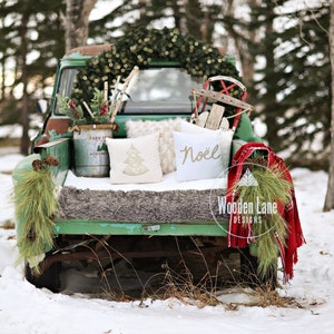 May include: A vintage green truck bed decorated for Christmas with a wreath, pillows, and a red blanket. The truck is parked in a snowy setting with trees in the background. The pillows have the words "Noël" and a Christmas tree design.