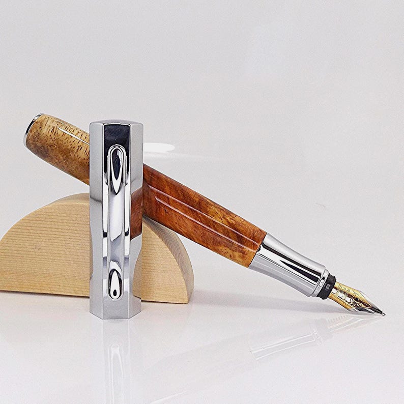 May include: A luxury fountain pen with a polished silver cap and a wooden barrel. The pen features a gold nib and is displayed on a wooden stand. The barrel has a rich, brown wood grain pattern.