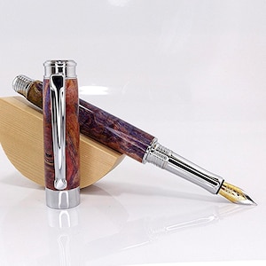 May include: A fountain pen with a wooden barrel in shades of red, purple, and brown. The pen has a silver-coloured metal clip and accents, and a gold-coloured nib. The pen is displayed with its cap off, resting on a wooden stand.