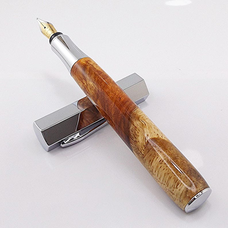 May include: A handcrafted fountain pen with a polished wood grain barrel and a silver-colored metal cap and accents. The pen features a gold-colored nib and rests on its cap, showcasing its elegant design. The pen is a writing instrument.