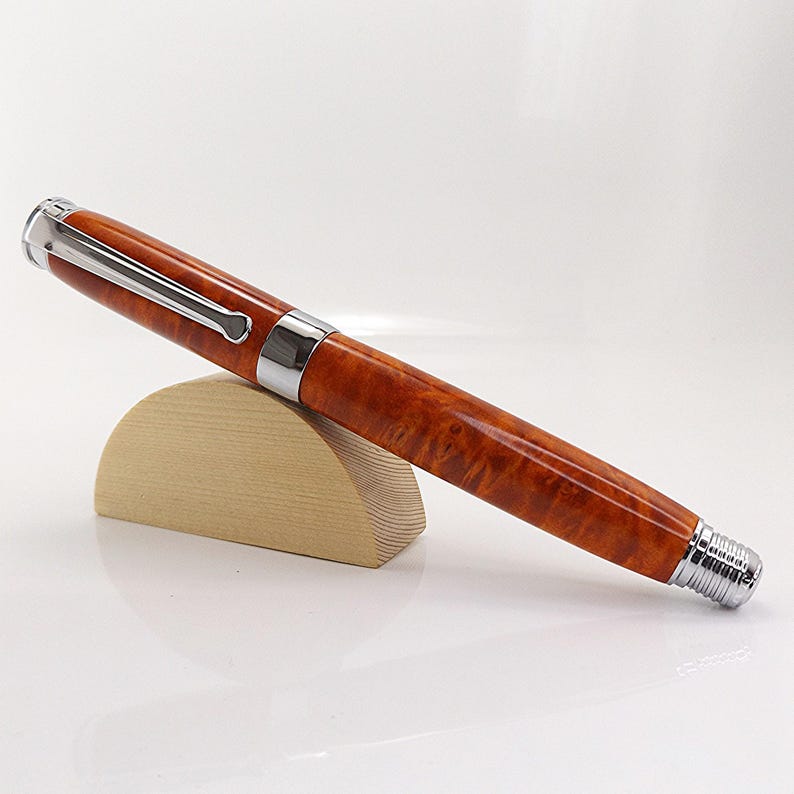 Op de afbeelding: Een luxe vulpen met een gepolijste, roodbruine houten schacht en zilverkleurige metalen accenten. De pen rust op een kleine, lichtgekleurde houten standaard, die het ontwerp laat zien. De dop en punt van de pen zijn ook zilverkleurig.