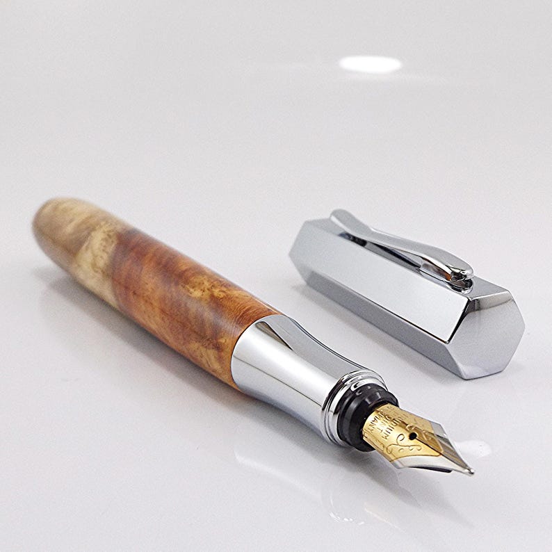May include: A fountain pen with a wooden barrel and a silver-colored cap and trim. The pen's barrel has a natural wood grain pattern in shades of brown and tan. The nib is gold-colored.