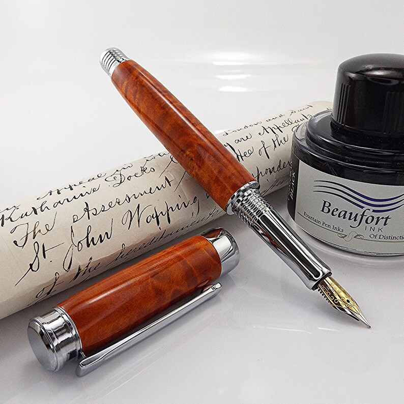 Op de afbeelding: Een houten vulpen met zilverkleurige metalen details, naast een fles zwarte inkt en een opgerold document. De dop van de pen is los. Op de inktfles staat "Beaufort Fountain Pen Inks". Het document heeft handgeschreven tekst.