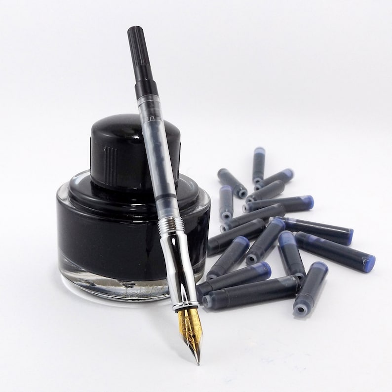 May include: A fountain pen with a gold nib, a clear barrel, and a black cap rests beside a black ink bottle and a scattering of ink cartridges. The ink is a deep black color, and the background is white.