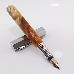 May include: A handcrafted fountain pen with a polished wood barrel and a silver-colored metal grip and cap. The pen features a gold-colored nib and rests on its silver cap. The wood grain has a natural, light brown and tan pattern.