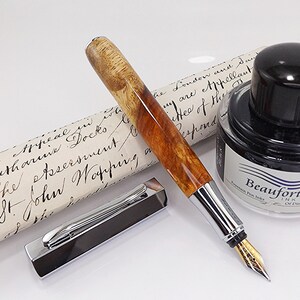 May include: A luxury fountain pen with a wooden barrel and silver accents, alongside a bottle of black ink and a historical document. The pen's nib is gold-colored, and the pen cap is detached.