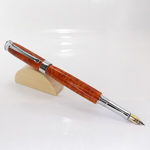 Op de afbeelding: Een luxe vulpen met een gepolijste, roodbruine houten schacht en zilverkleurige metalen accenten. De pen heeft een goudkleurige penpunt en rust op een kleine, lichtgekleurde houten standaard. De pen is ontworpen om mee te schrijven.