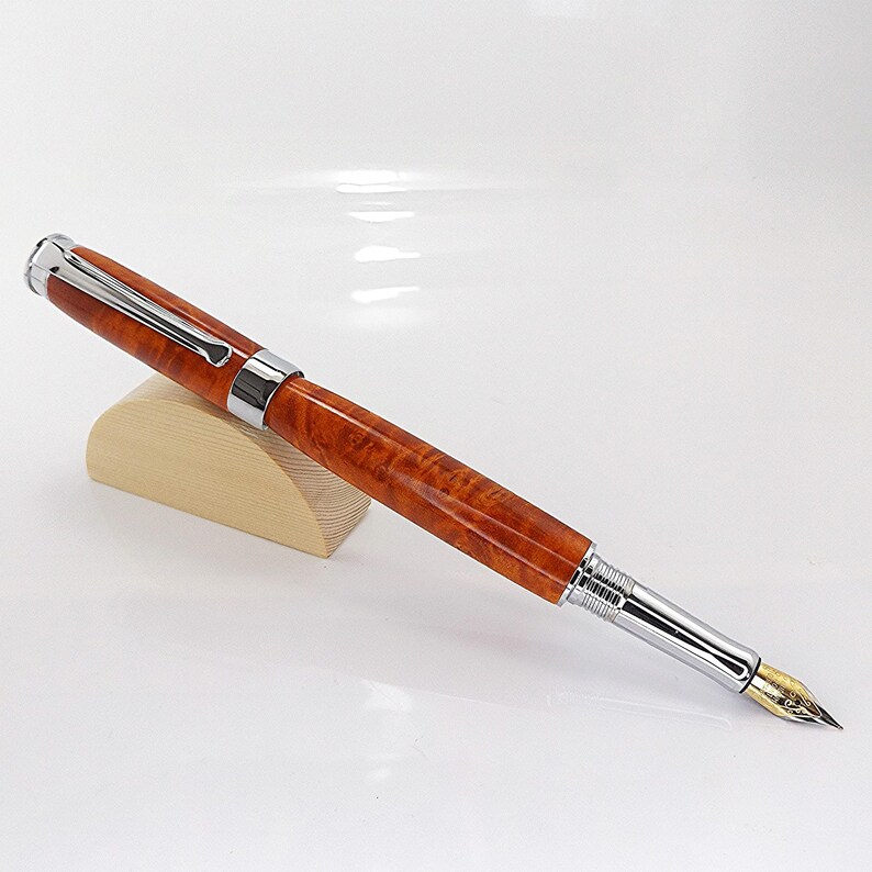 Op de afbeelding: Een luxe vulpen met een gepolijste, roodbruine houten schacht en zilverkleurige metalen accenten. De pen heeft een goudkleurige penpunt en rust op een kleine, lichtgekleurde houten standaard. De pen is ontworpen om mee te schrijven.