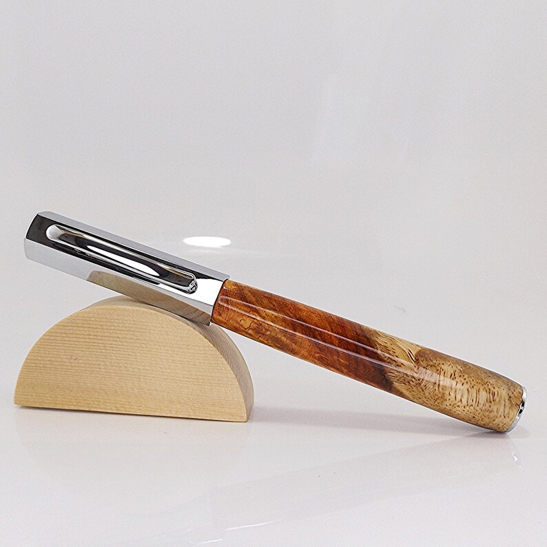 May include: A handcrafted pen with a polished chrome cap and a wooden barrel. The barrel features a natural wood grain pattern in shades of brown and tan. The pen rests on a small, light-colored wooden stand.