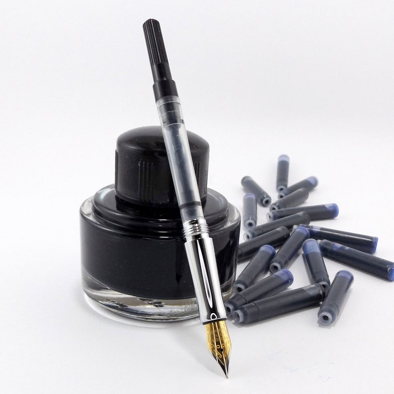 Op de afbeelding: Een vulpen met een gouden penpunt ligt naast een zwarte inktfles en verschillende inktpatronen. De pen heeft een doorzichtige schacht en de inktfles is ovaal van vorm. De patronen zijn zwart met blauwe uiteinden.