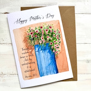Pode incluir: Um cartão de aquarela do Dia das Mães com um arranjo floral em um vaso azul. O cartão apresenta o texto "Happy Mother's Day" e um versículo bíblico. Um envelope marrom está atrás do cartão.