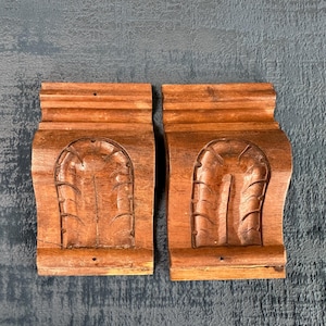 Peut inclure: Deux corbeaux en bois avec un motif de feuille sculpté. Les corbeaux sont de couleur brun clair et ont une forme légèrement incurvée.