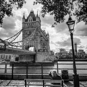 May include: A monochrome image of Tower Bridge in London. The bridge's towers and walkways are prominent. A person sits on a bench, and a traditional lamppost stands nearby. The sky is overcast.