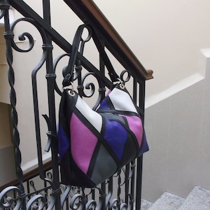 Könnte beinhalten: Eine bunte Patchwork-Handtasche mit schwarzen, weißen, violetten und rosafarbenen Lederpaneelen. Die Tasche hängt an einem schwarzen Metallgeländer.