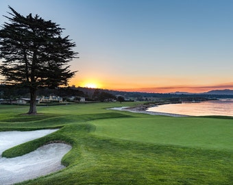 Puede incluir: Un campo de golf al atardecer. La imagen presenta una calle verde, una trampa de arena y una bandera en el green. Un gran árbol se alza a la izquierda, con una masa de agua y un cielo colorido al fondo.