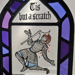 Puede incluir: Panel de vidriera con una ilustración medieval de un caballero herido y el texto "Tis but a scratch". El panel tiene un marco negro con detalles de vidrio morado y azul. El caballero tiene una herida sangrienta en la cabeza.