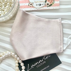 Puede incluir: Una mascarilla facial rosa claro con lazos el&aacute;sticos blancos para las orejas y un acento de perla en las ataduras. La mascarilla est&aacute; hecha de una tela suave y sedosa. El texto "Lamartei Couture" es visible en una tarjeta negra en la imagen.