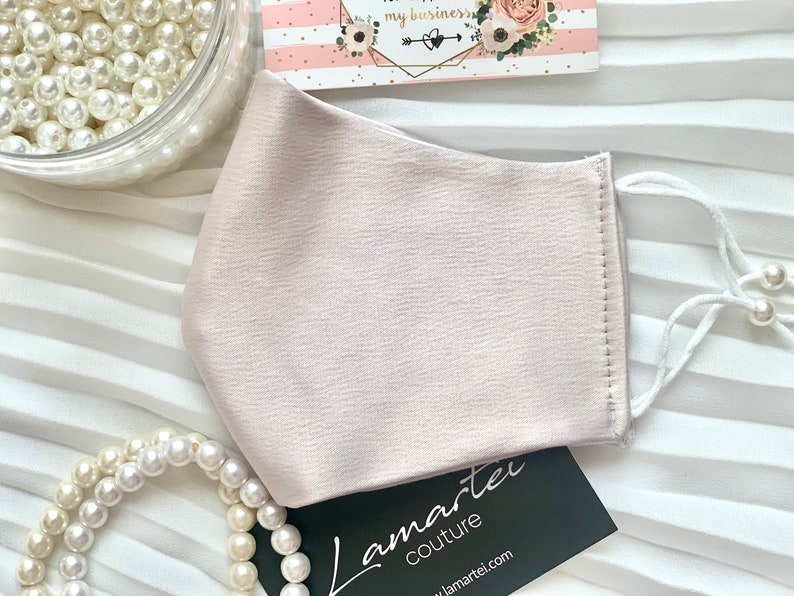 Puede incluir: Una mascarilla facial rosa claro con lazos el&aacute;sticos blancos para las orejas y un acento de perla en las ataduras. La mascarilla est&aacute; hecha de una tela suave y sedosa. El texto "Lamartei Couture" es visible en una tarjeta negra en la imagen.