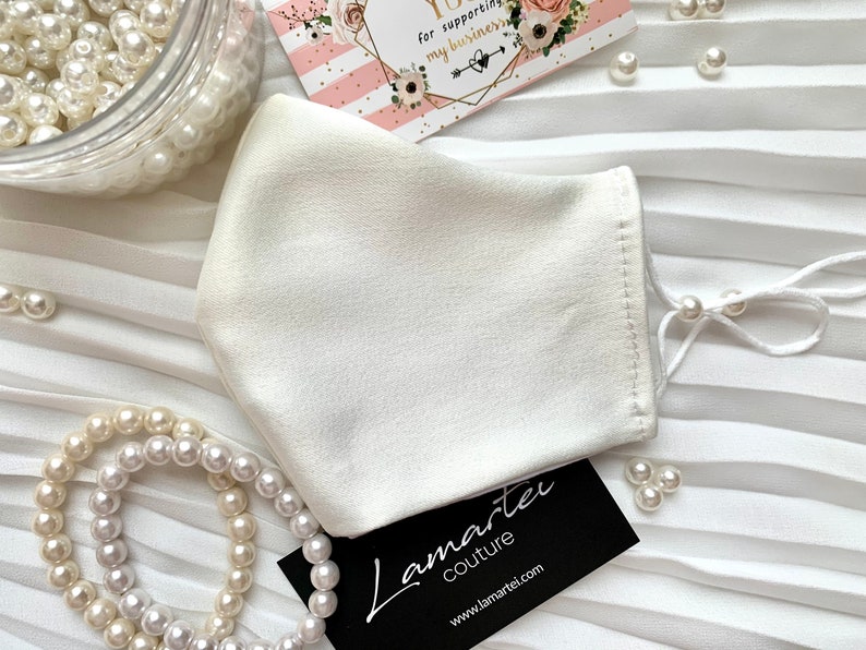 Puede incluir: M&aacute;scara facial blanca de sat&eacute;n con el&aacute;sticos de oreja blancos y cuentas de perlas blancas. La m&aacute;scara est&aacute; sobre una superficie de tela blanca plisada. Una tarjeta de visita con el texto "Lamartei Couture" y la direcci&oacute;n del sitio web "www.lamartei.com" est&aacute; en la esquina inferior derecha de la imagen.
