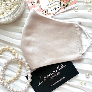 Puede incluir: Una mascarilla facial beige claro de sat&eacute;n con el&aacute;sticos de oreja blancos. La mascarilla est&aacute; sobre un fondo blanco con cuentas de perlas y una tarjeta de visita que dice "Lamartei Couture" y "www.lamartei.com".