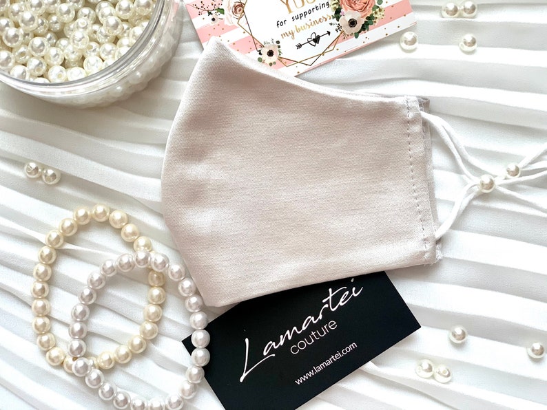Puede incluir: Una mascarilla facial beige claro de sat&eacute;n con el&aacute;sticos de oreja blancos. La mascarilla est&aacute; sobre un fondo blanco con cuentas de perlas y una tarjeta de visita que dice "Lamartei Couture" y "www.lamartei.com".
