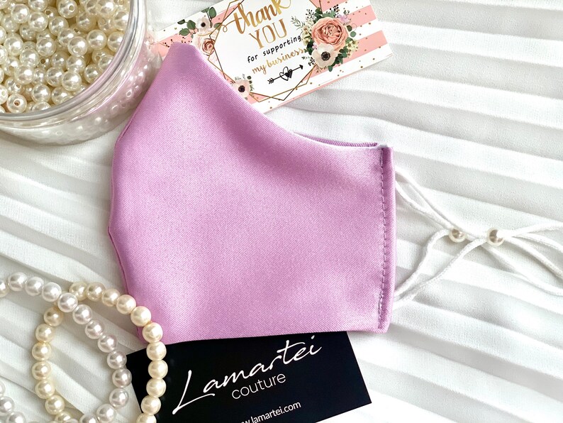 Puede incluir: Una mascarilla facial de sat&eacute;n de color lila claro con lazos blancos y cuentas blancas. La mascarilla est&aacute; sobre una superficie blanca con una tarjeta de visita que dice "Lamartei Couture" y una tarjeta de agradecimiento que dice "Gracias por apoyar mi negocio".