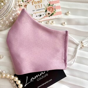 Puede incluir: Una mascarilla facial de color lila claro hecha de tejido satinado con correas ajustables blancas y cuentas de perlas blancas. La mascarilla est&aacute; sobre una superficie blanca con una tarjeta de visita que dice "Lamari Couture" y una tarjeta de agradecimiento.