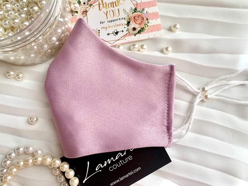 Puede incluir: Una mascarilla facial de color lila claro hecha de tejido satinado con correas ajustables blancas y cuentas de perlas blancas. La mascarilla est&aacute; sobre una superficie blanca con una tarjeta de visita que dice "Lamari Couture" y una tarjeta de agradecimiento.