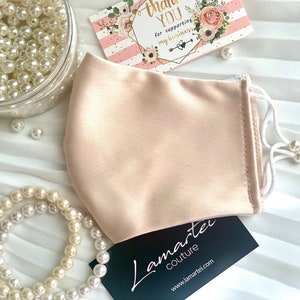 Puede incluir: Una mascarilla facial de sat&eacute;n rosa claro con el&aacute;sticos de oreja blancos. La mascarilla est&aacute; sobre una superficie blanca con perlas y una tarjeta de agradecimiento.
