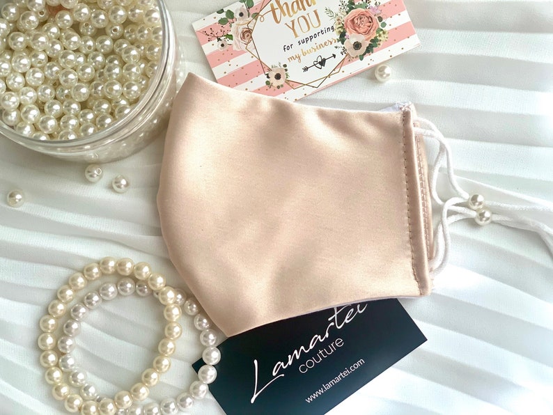 Puede incluir: Una mascarilla facial de sat&eacute;n rosa claro con el&aacute;sticos de oreja blancos. La mascarilla est&aacute; sobre una superficie blanca con perlas y una tarjeta de agradecimiento.