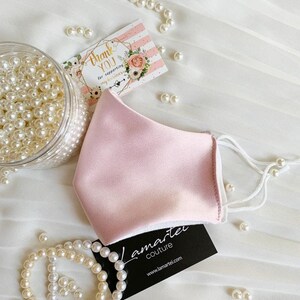 Puede incluir: Una mascarilla facial de sat&eacute;n rosa con el&aacute;sticos de oreja blancos. La mascarilla est&aacute; sobre una superficie blanca con cuentas de perlas y una tarjeta de visita que dice "Lamartei Couture" y "www.lamartei.com".