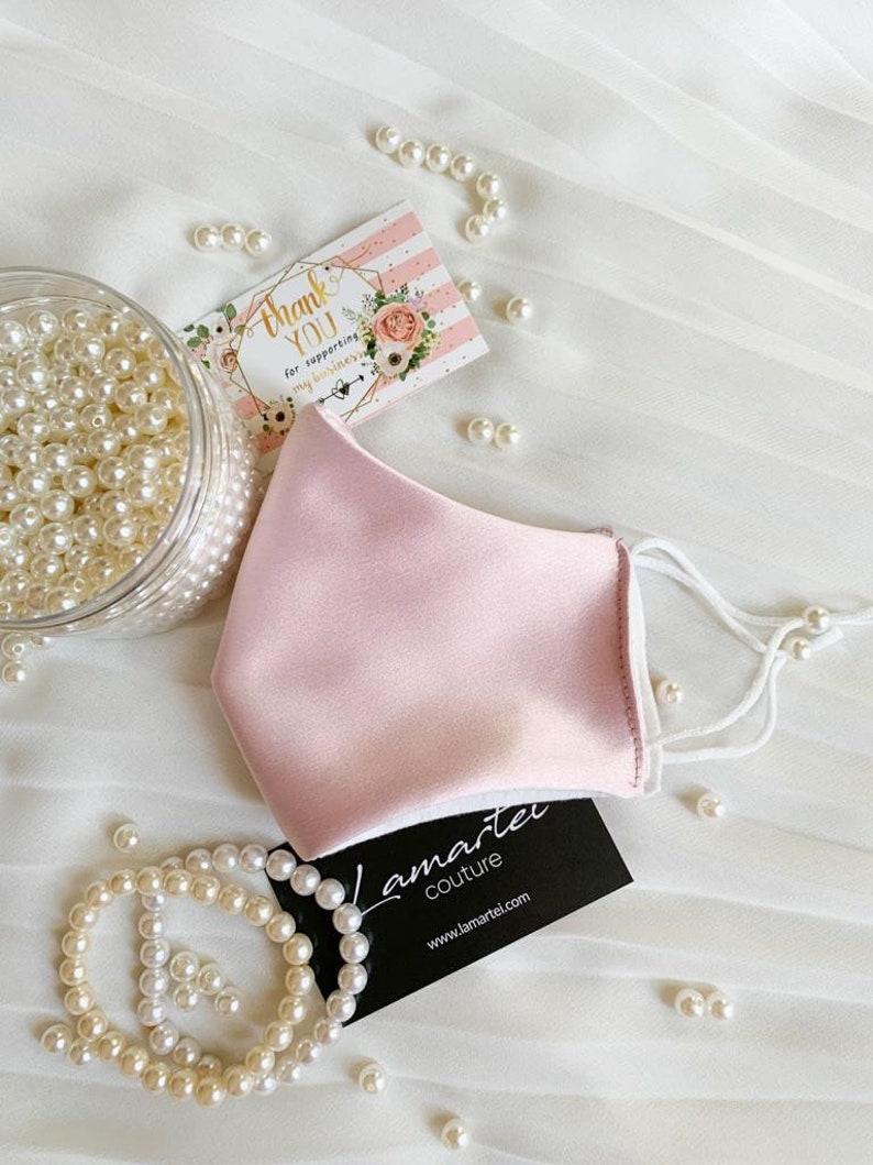 Puede incluir: Una mascarilla facial de sat&eacute;n rosa con el&aacute;sticos de oreja blancos. La mascarilla est&aacute; sobre una superficie blanca con cuentas de perlas y una tarjeta de visita que dice "Lamartei Couture" y "www.lamartei.com".