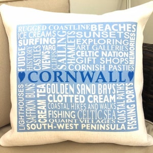 May include: A white pillow with a blue square design featuring the word "Cornwall" in blue lettering surrounded by a word cloud of things to do and see in Cornwall, England. The words include "beaches", "ice creams", "surfing", "yarg", "exploring", "art galleries", "Celtic nation", "gift shops", "Cornish pasties", "lighthouses", "saffron buns", "cream tea", "golden sand bays", "clotted cream", "coastal hikes and walks", "fishing", "Celtic sea", "quaint villages", "south-west peninsula", and "fishing ports".