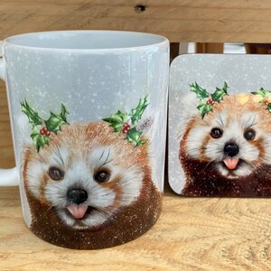 May include: A white ceramic mug and square coaster set. The design features a red panda wearing a holly wreath, with its tongue out. The background is light blue with white snowflakes.