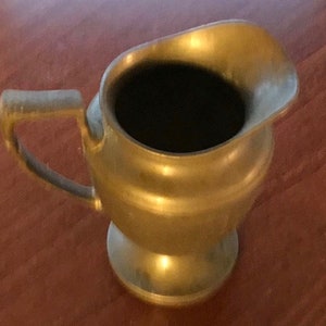 May include: A gold metal pitcher with a rounded body and a handle. The pitcher has a smooth, polished finish.