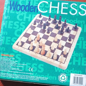 Può includere: Un set di scacchi in legno in una scatola verde e bianca. La scatola ha le parole "Wooden Chess" stampate in grandi caratteri grassetto. La scacchiera è fatta di legno e ha un colore marrone chiaro. I pezzi degli scacchi sono anch'essi fatti di legno e sono dipinti di nero e bianco. La scatola include anche le parole "SIMPLEX" e "SX70000".