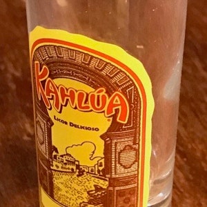 May include: A clear glass shot glass with a yellow and red label that says "Kahlua Licor Delicioso Liqueur". The label has a brown illustration of a building with a yellow archway.